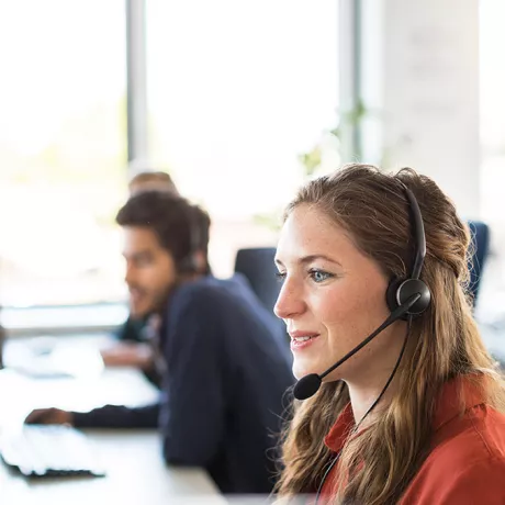 Wsparcie klienta Foto einer Frau mit Headset, die in einem Großraumbüro sitzt und für Kundensupport zuständig ist